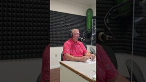 A man in a bright pink shirt, seated in a soundproof studio, speaks into a microphone. He wears headphones and appears focused, with acoustic panels visible in the background.