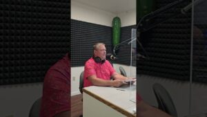 A man wearing a red shirt and headphones sits at a desk with a microphone in a soundproof room. He appears focused, with acoustic foam panels around him.