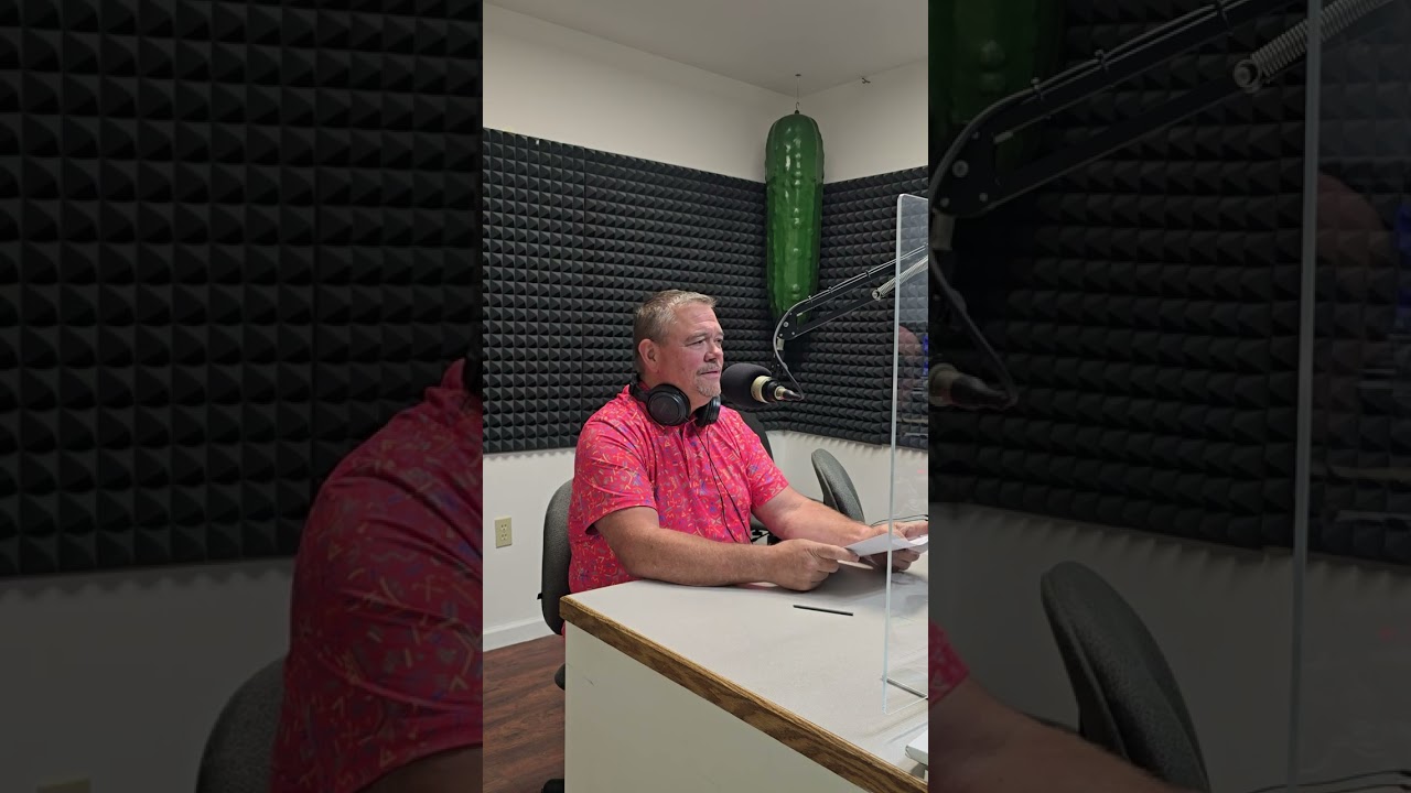 A man wearing a red shirt and headphones sits at a desk with a microphone in a soundproof room. He appears focused, with acoustic foam panels around him.