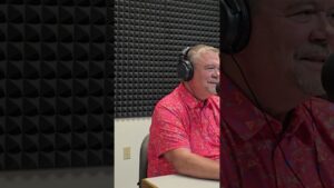 Man in a vibrant pink shirt with geometric patterns sits at a recording studio desk, wearing headphones, surrounded by foam-panel walls, smiling.