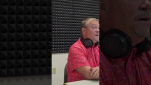 A man in a patterned pink shirt is sitting in a studio with soundproof foam walls. He has headphones resting around his neck and appears to be speaking.