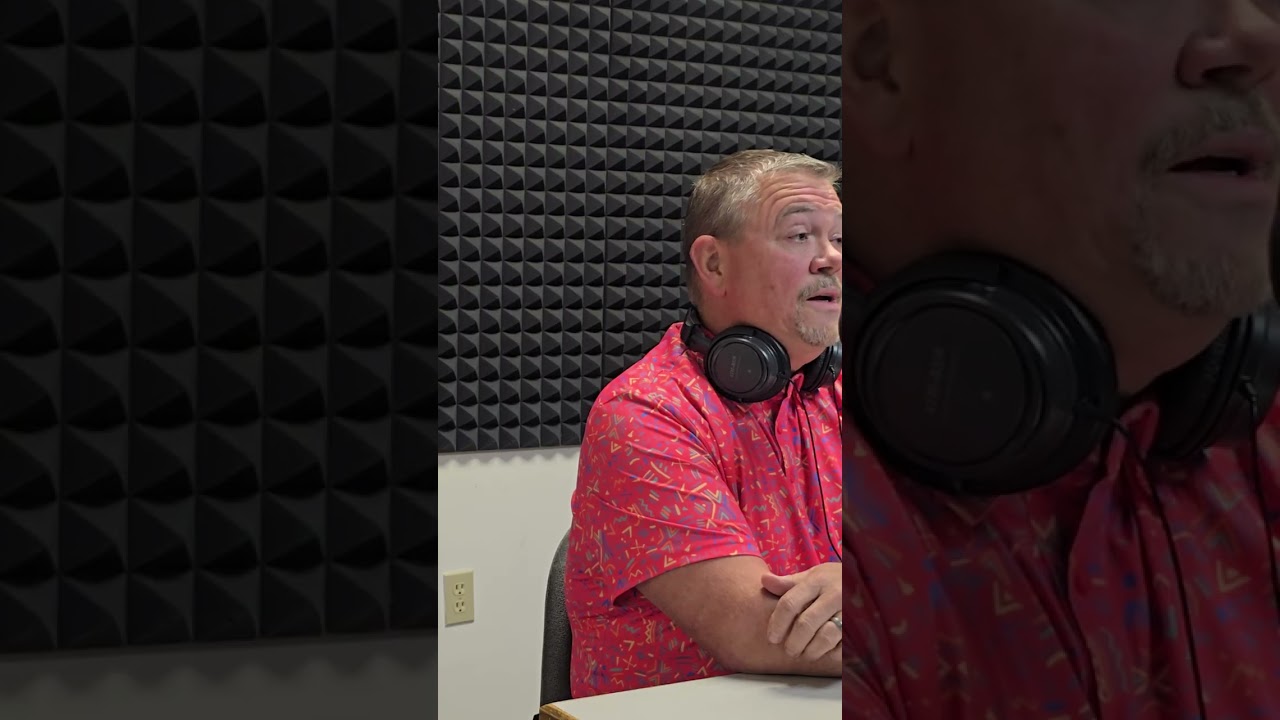 A man in a patterned pink shirt is sitting in a studio with soundproof foam walls. He has headphones resting around his neck and appears to be speaking.